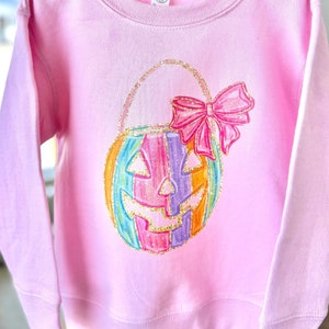 Puede incluir: Una sudadera rosa con una calabaza rayada arco&iacute;ris con un lazo rosa en la parte superior. La calabaza es una bolsa de truco o trato.
