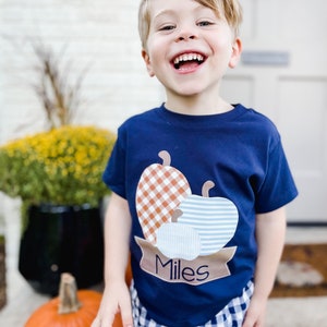 May include: A young boy wearing a navy blue t-shirt with a patch of three pumpkins in orange, brown, and blue plaid. The pumpkins are on a brown banner with the name "Miles" written in black.