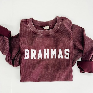 May include: A maroon long-sleeved shirt with a white ribbed texture and the word "BRAHMAS" printed in white on the front.