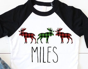 Op de afbeelding: Een wit en zwart shirt met lange mouwen met een rood en groen geruit eland ontwerp en de naam "MILES" gedrukt op de voorkant. Het shirt wordt gedragen door een kindmodel dat blauwe jeans en witte sneakers draagt.