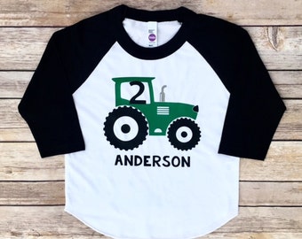 Puede incluir: Camiseta de béisbol blanca y negra con un gráfico de tractor verde y el número 2 en el lateral. El texto "ANDERSON" está impreso debajo del tractor.