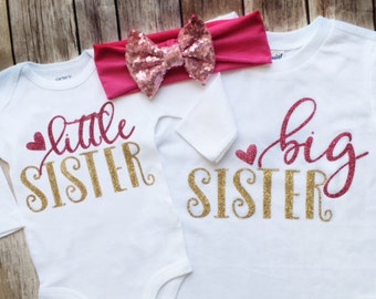 May include: Two white shirts with gold glitter lettering. One shirt says "little sister" with a pink heart and the other says "big sister" with a pink heart. A pink headband with a sequined bow is included.