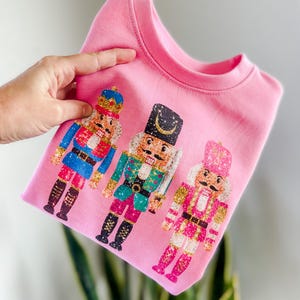 Può includere: Felpa rosa con una stampa glitterata di tre soldati di noce. I soldati di noce indossano costumi colorati e hanno accenti dorati.