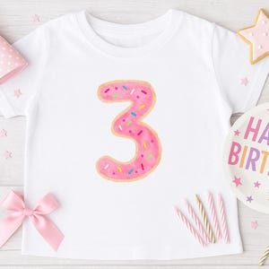 Op de afbeelding: Wit T-shirt met een roze donutvormig nummer 3 met kleurrijke sprinkels.