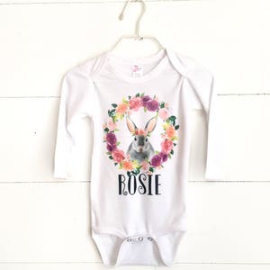 May include: White long-sleeved baby onesie with a floral wreath and a bunny wearing a flower crown. The onesie has the name "Rosie" printed on the front.