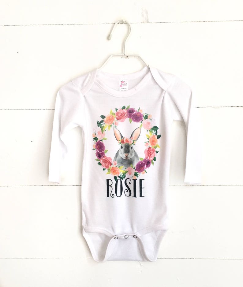 May include: White long-sleeved baby onesie with a floral wreath and a bunny wearing a flower crown. The onesie has the name "Rosie" printed on the front.