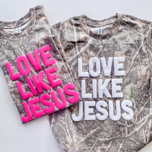 May include: Two camouflage-patterned t-shirts. One shirt has the words "LOVE LIKE JESUS" in bright pink letters, and the other has the same phrase in white letters. The shirts are made of a soft material.