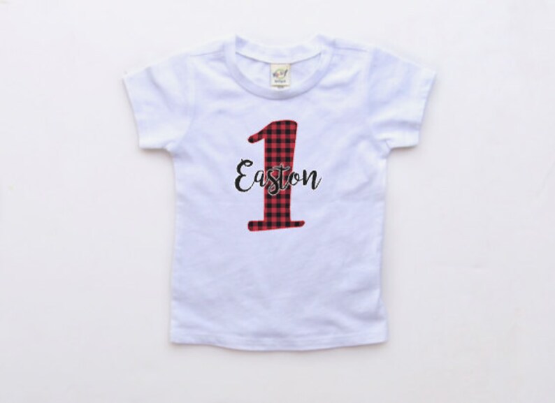 May include: White short-sleeved t-shirt with a red and black plaid number one and the name Easton in black script.