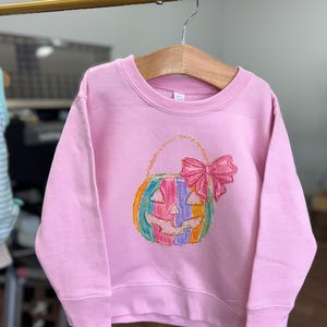 Puede incluir: Una sudadera rosa con una calabaza rayada arco&iacute;ris con un lazo rosa en la parte superior. La calabaza es una bolsa de truco o trato.
