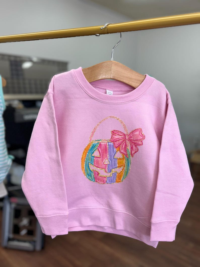 Puede incluir: Una sudadera rosa con una calabaza rayada arco&iacute;ris con un lazo rosa en la parte superior. La calabaza es una bolsa de truco o trato.