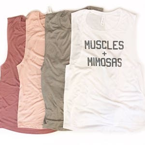 Puede incluir: Cuatro camisetas sin mangas de diferentes colores. La camiseta blanca tiene el texto "MUSCLES + MIMOSAS" impreso.