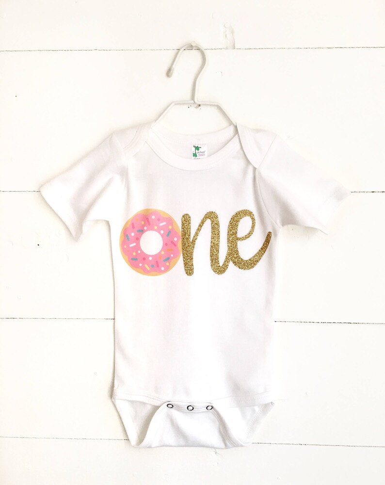 Pode incluir: Macac&atilde;o branco com um gr&aacute;fico de donut rosa e marrom e texto de glitter dourado que diz "one".