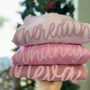 以下が含まれることがあります： ピンクのトレーナーが3枚重ねられています。トレーナーには、「Meredith」、「Whitney」、「Leila」という名前がピンクの文字で書かれています。