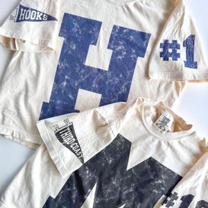 Puede incluir: Dos camisetas de color blanco roto con gráficos azul marino desgastados. La superior tiene una letra H grande, un banderín en la manga con el texto HOOKS y #1. La inferior tiene una letra M grande, un banderín con THIRD COAST y #19.