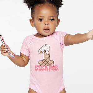 May include: Pink baby onesie with a graphic of an ice cream cone shaped like the number one. The text "Eleanor" is printed below the ice cream cone.