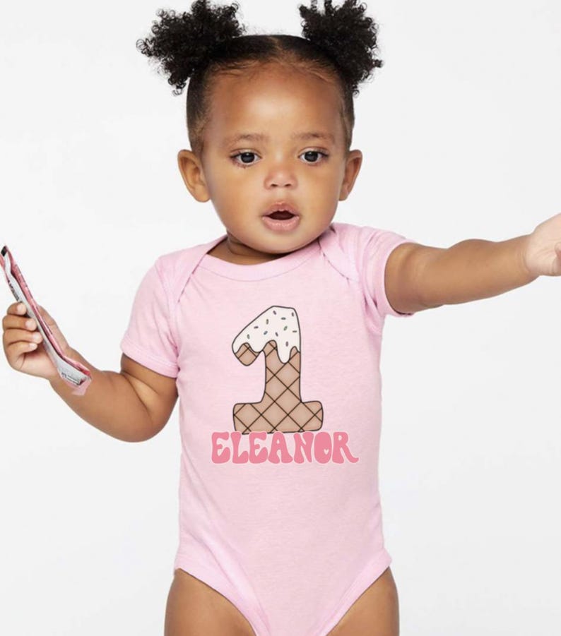 May include: Pink baby onesie with a graphic of an ice cream cone shaped like the number one. The text "Eleanor" is printed below the ice cream cone.
