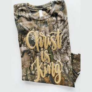 May include: A folded camouflage-patterned t-shirt with the words "Christ is King" in gold script. The shirt has a crew neck and is made of a soft, likely cotton, material. The camouflage design features shades of brown, green, and gray.