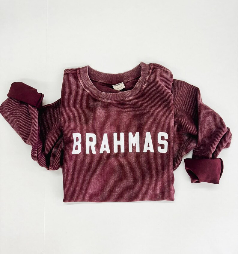 May include: A maroon long-sleeved shirt with a white ribbed texture and the word "BRAHMAS" printed in white on the front.