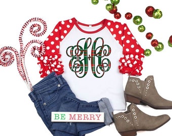 Op de afbeelding: Een wit raglan shirt met rode stippen mouwen en een groen en rood geruit monogram. Het shirt wordt gecombineerd met een spijkerbroek en grijze enkellaarsjes. De zin "BE MERRY" staat op een klein houten bord.