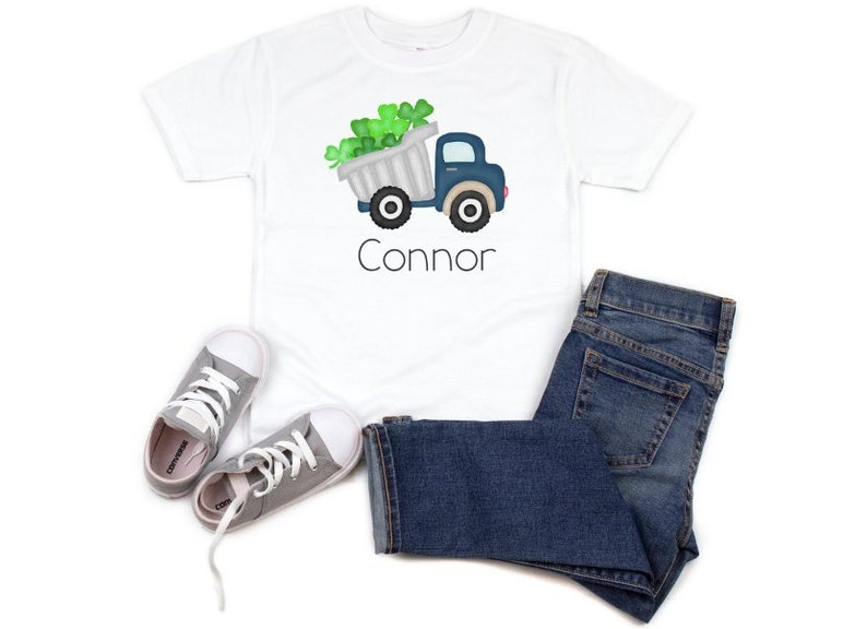 May include: White t-shirt with a blue truck carrying a load of green shamrocks. The truck has the name "Connor" printed below it.