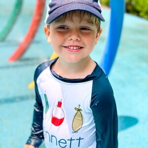 May include: A young boy wearing a blue and white baseball cap with a red and white design on the front. He is wearing a white long-sleeved shirt with a black and blue design on the sleeves. The shirt has a red and white fishing bobber, a green fishing lure, and a yellow and white fishing lure on the front. The shirt also has the word "Bennett" printed on the front.