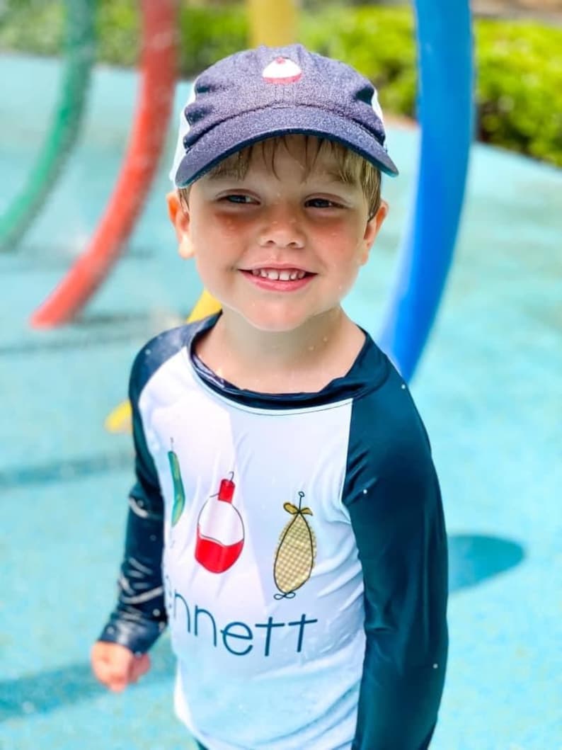 May include: A young boy wearing a blue and white baseball cap with a red and white design on the front. He is wearing a white long-sleeved shirt with a black and blue design on the sleeves. The shirt has a red and white fishing bobber, a green fishing lure, and a yellow and white fishing lure on the front. The shirt also has the word "Bennett" printed on the front.