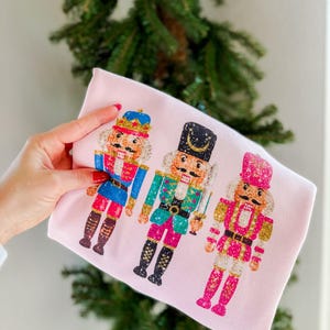 May include: Pink long-sleeved shirt with a graphic of three nutcracker soldiers. The nutcrackers are wearing colorful, glittery outfits and have gold accents.