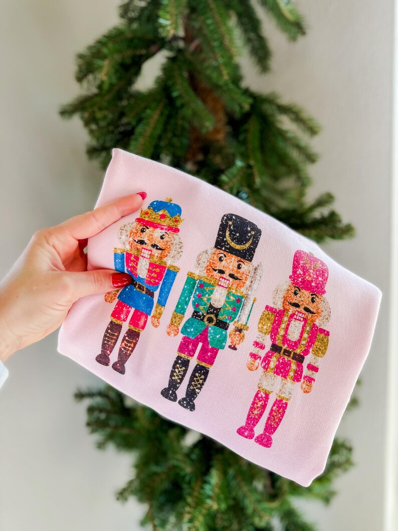 May include: Pink long-sleeved shirt with a graphic of three nutcracker soldiers. The nutcrackers are wearing colorful, glittery outfits and have gold accents.