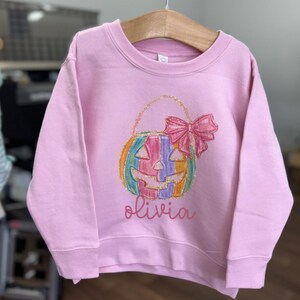 Puede incluir: Sudadera rosa de manga larga con una calabaza arco&iacute;ris que lleva un lazo y el nombre "olivia" impreso en la parte delantera.