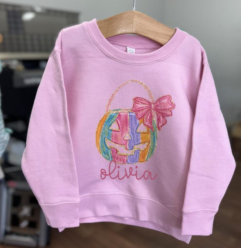Puede incluir: Sudadera rosa de manga larga con una calabaza arco&iacute;ris que lleva un lazo y el nombre "olivia" impreso en la parte delantera.