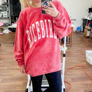 May include: A woman wearing a pink corduroy sweatshirt with the word "RICEBIRD" in white lettering. She is also wearing black leggings and white sneakers.