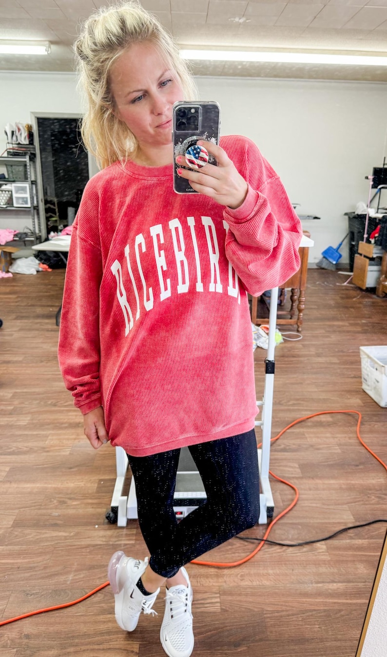 May include: A woman wearing a pink corduroy sweatshirt with the word "RICEBIRD" in white lettering. She is also wearing black leggings and white sneakers.