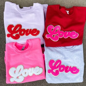 May include: Four sweatshirts in different colors with the word "Love" in a retro font on the front. The sweatshirts are in pink, white, red, and gray.