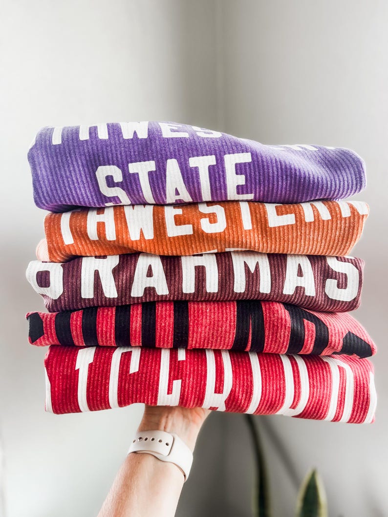 May include: A stack of five folded long-sleeved shirts with different colors and text. The top shirt is purple with the text "STATE" in white. The second shirt is orange with the text "AWESOM" in white. The third shirt is brown with the text "GRAMMAS" in white. The fourth shirt is red with black stripes and the text "I LOVE" in white. The bottom shirt is red with the text "TCU" in white.