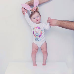 May include: A baby girl wearing a white long-sleeved onesie with a floral wreath and bunny design. The onesie has the name "Rosie" printed on it. The baby is wearing pink bunny ears and is standing on a white surface.