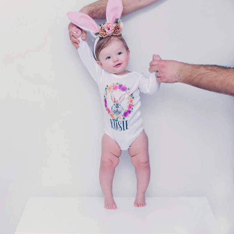 May include: A baby girl wearing a white long-sleeved onesie with a floral wreath and bunny design. The onesie has the name "Rosie" printed on it. The baby is wearing pink bunny ears and is standing on a white surface.