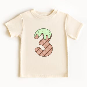 May include: A cream colored t-shirt with a green and brown ice cream cone shaped number three.