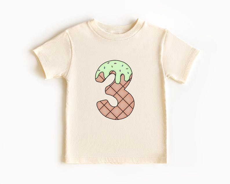 May include: A cream colored t-shirt with a green and brown ice cream cone shaped number three.