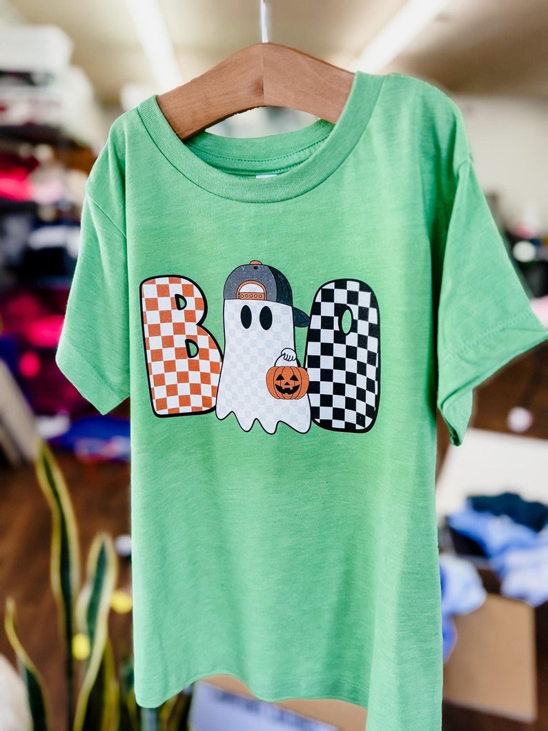 May include: Green short-sleeved t-shirt with a checkered "BOO" graphic featuring a ghost wearing a baseball cap and holding a pumpkin.