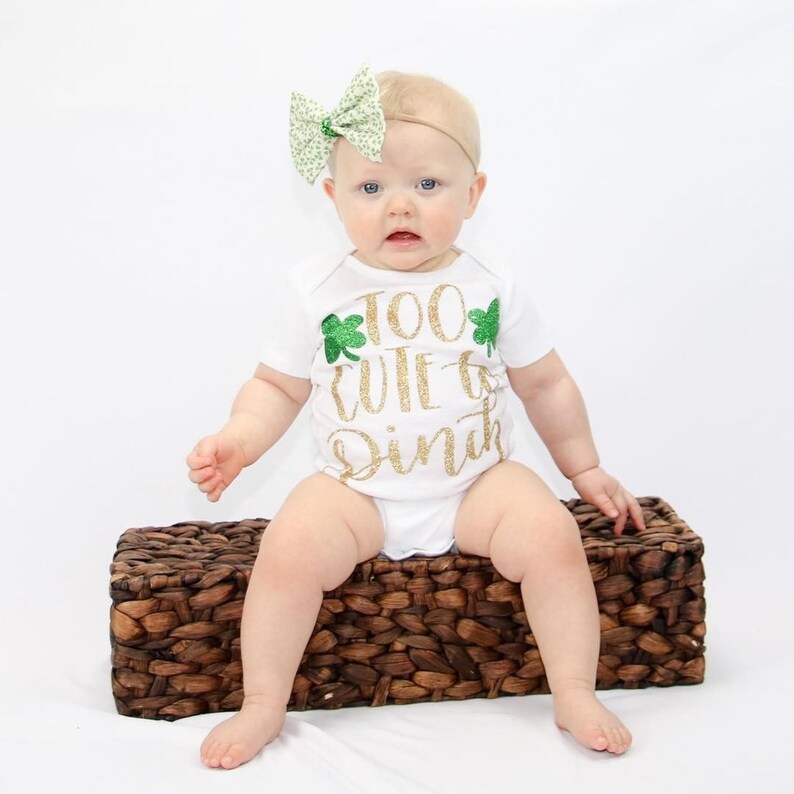 May include: A baby wearing a white onesie with gold glitter text that reads "Too cute to pinch" and green shamrock accents. The baby is wearing a green and white bow headband and is sitting on a brown wicker basket.