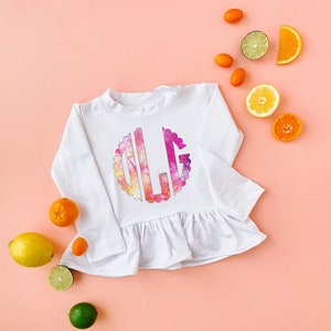 Pode incluir: Camisola branca de manga comprida com bainha franzida e um grande monograma "LLG" em tie-dye colorido na frente.