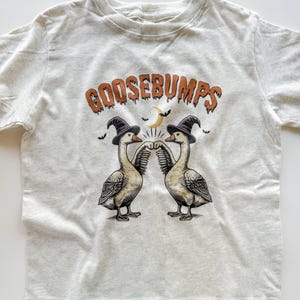 May include: A light gray t-shirt featuring the word "GOOSEBUMPS" in orange lettering. Two geese wearing witch hats are fist-bumping. A crescent moon and bats are also featured in the design. The shirt is made of a soft material.