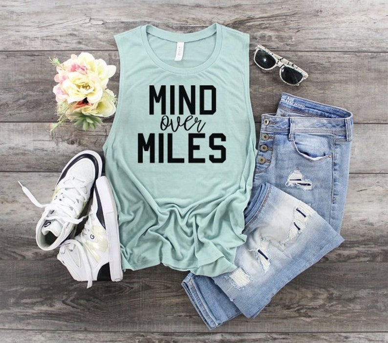 Pu&ograve; includere: Un top senza maniche di colore azzurro chiaro con la scritta "MIND over MILES" stampata in nero. Il top &egrave; appoggiato su una superficie di legno con un paio di scarpe da ginnastica bianche, un paio di jeans blu e un paio di occhiali da sole.