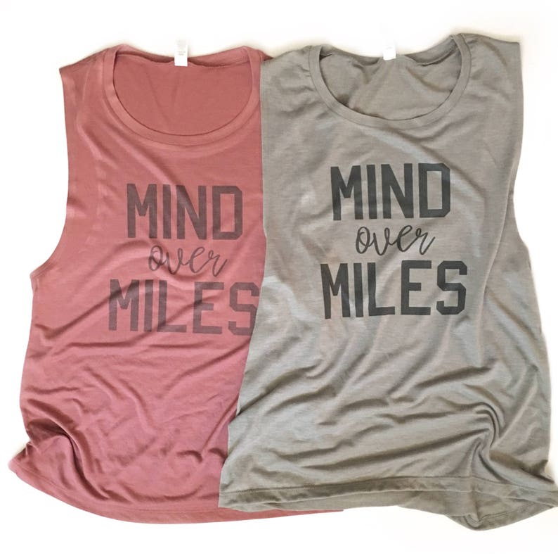Puede incluir: Dos camisetas sin mangas con el texto "Mind over Miles" impreso en ellas. Una camiseta es de color rosa polvoriento y la otra es gris claro.