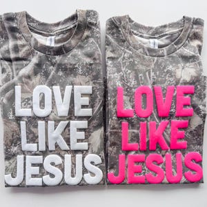 May include: Two short-sleeved t-shirts with a camouflage pattern in shades of brown, gray, and green. The left shirt features the text LOVE LIKE JESUS in raised white lettering, while the right shirt features the same text in raised neon pink.