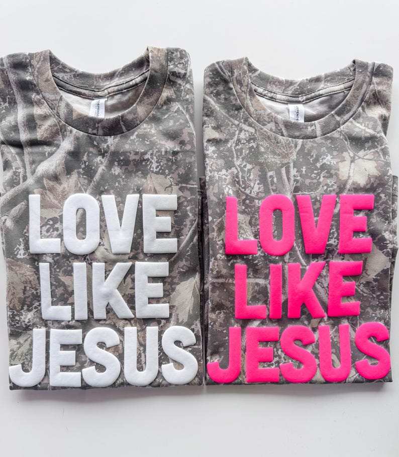 May include: Two short-sleeved t-shirts with a camouflage pattern in shades of brown, gray, and green. The left shirt features the text LOVE LIKE JESUS in raised white lettering, while the right shirt features the same text in raised neon pink.