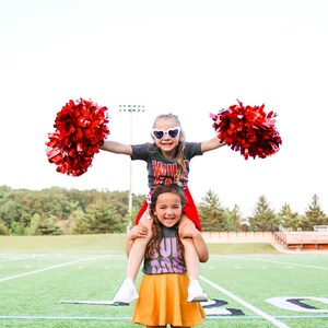K&ouml;nnte beinhalten: Zwei junge M&auml;dchen in Cheerleader-Outfits mit roten Pompons. Das M&auml;dchen vorne tr&auml;gt ein graues Shirt mit "Bulldog" in lila Buchstaben und einen gelben Rock. Das M&auml;dchen hinten tr&auml;gt ein graues Shirt mit "Wildcats" in roten Buchstaben und einen roten Rock. Sie stehen auf einem gr&uuml;nen Fu&szlig;ballfeld.