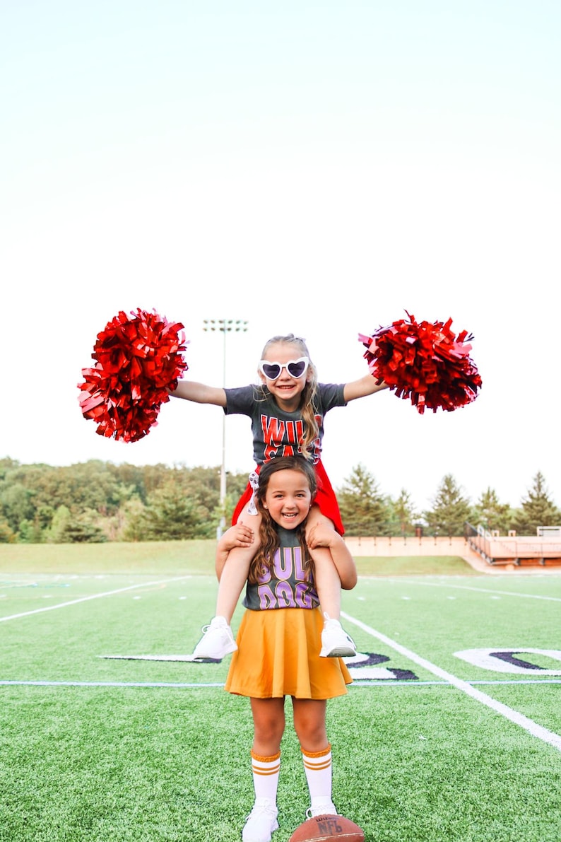K&ouml;nnte beinhalten: Zwei junge M&auml;dchen in Cheerleader-Outfits mit roten Pompons. Das M&auml;dchen vorne tr&auml;gt ein graues Shirt mit "Bulldog" in lila Buchstaben und einen gelben Rock. Das M&auml;dchen hinten tr&auml;gt ein graues Shirt mit "Wildcats" in roten Buchstaben und einen roten Rock. Sie stehen auf einem gr&uuml;nen Fu&szlig;ballfeld.