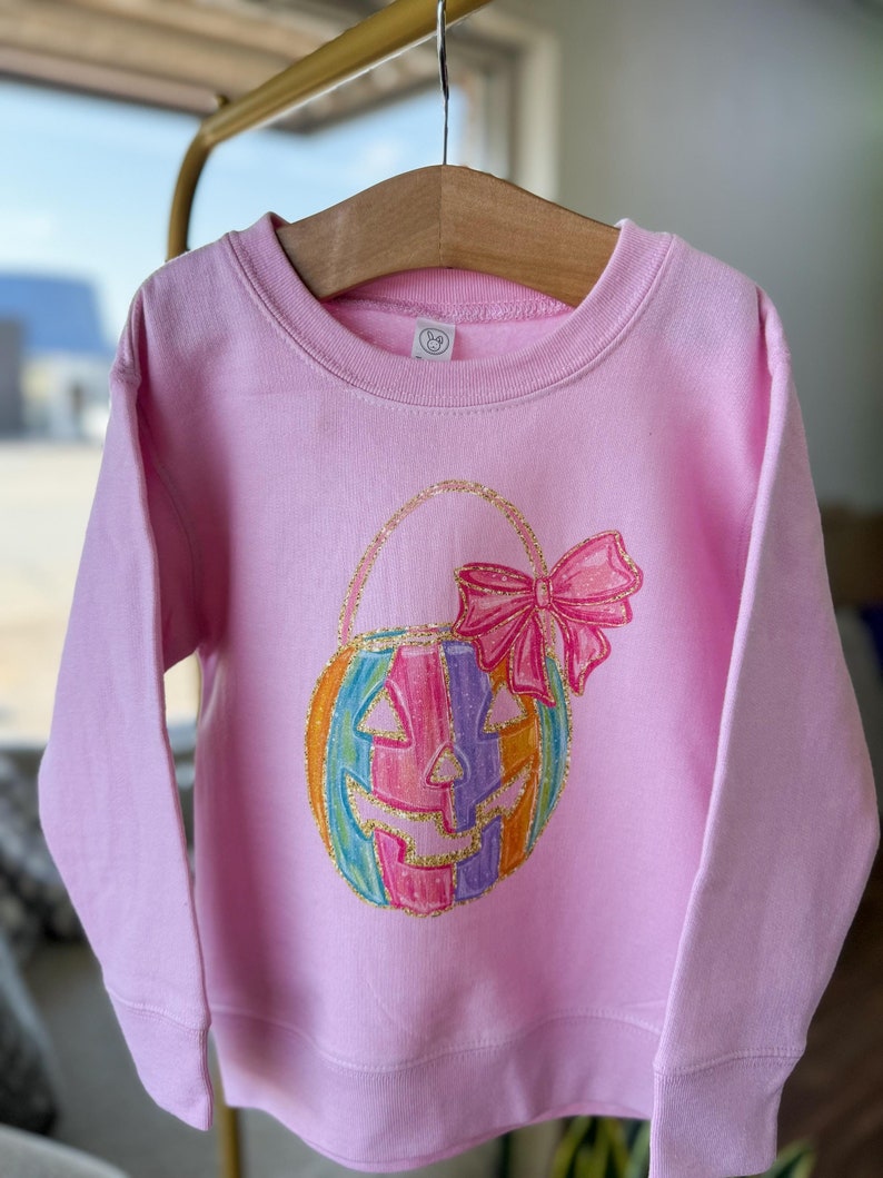 Puede incluir: Una sudadera rosa con un dise&ntilde;o de calabaza arco&iacute;ris con purpurina y un lazo rosa. La calabaza tiene una cara de jack-o'-lantern.
