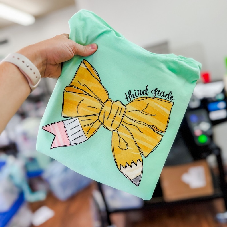 May include: A light green t-shirt with a yellow pencil bow and the text "third grade" in black script.